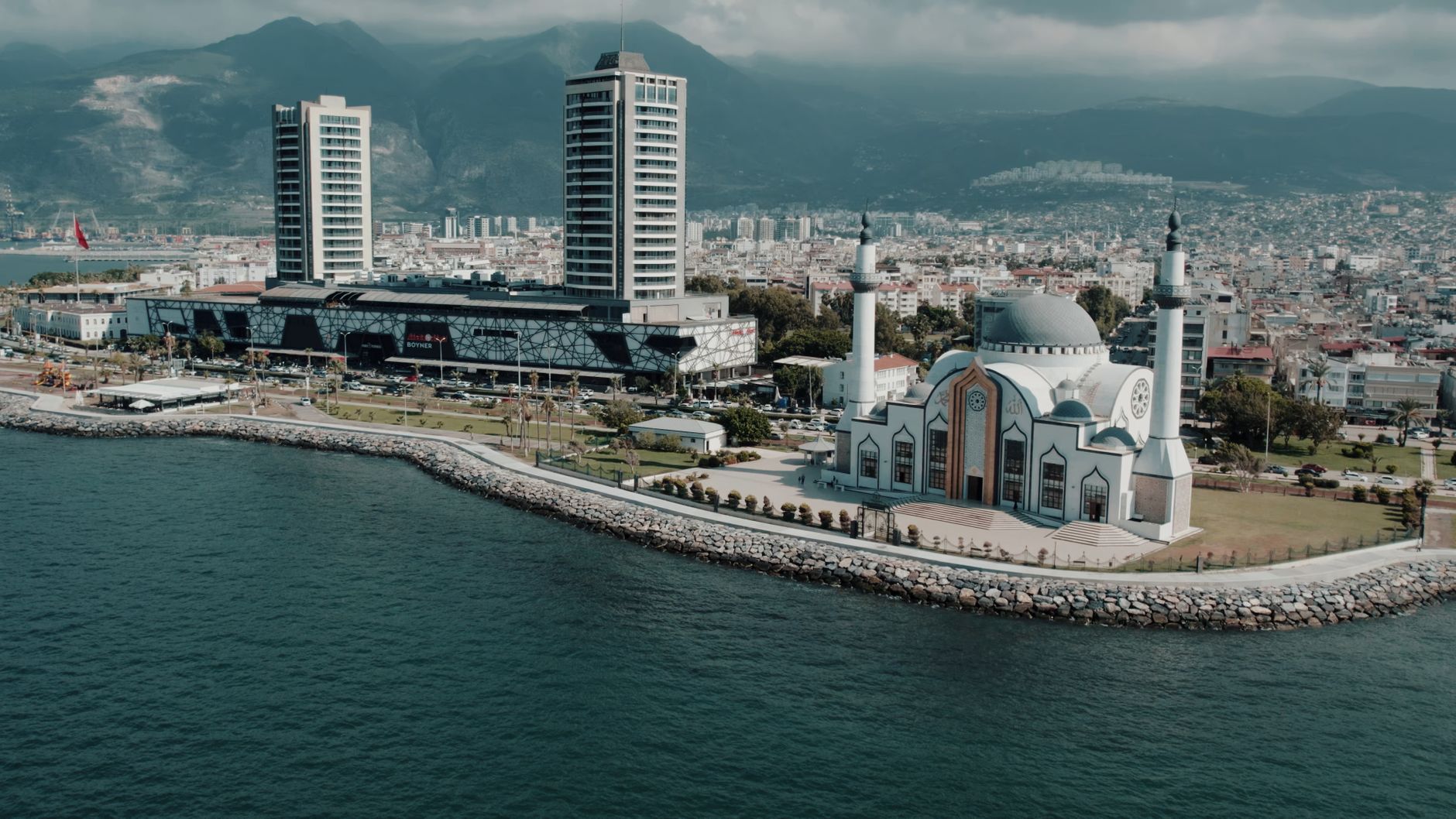 Aerial photograph showcasing İskenderun's mosque and cityscape against the mountainous backdrop on a sunny day. — Dar